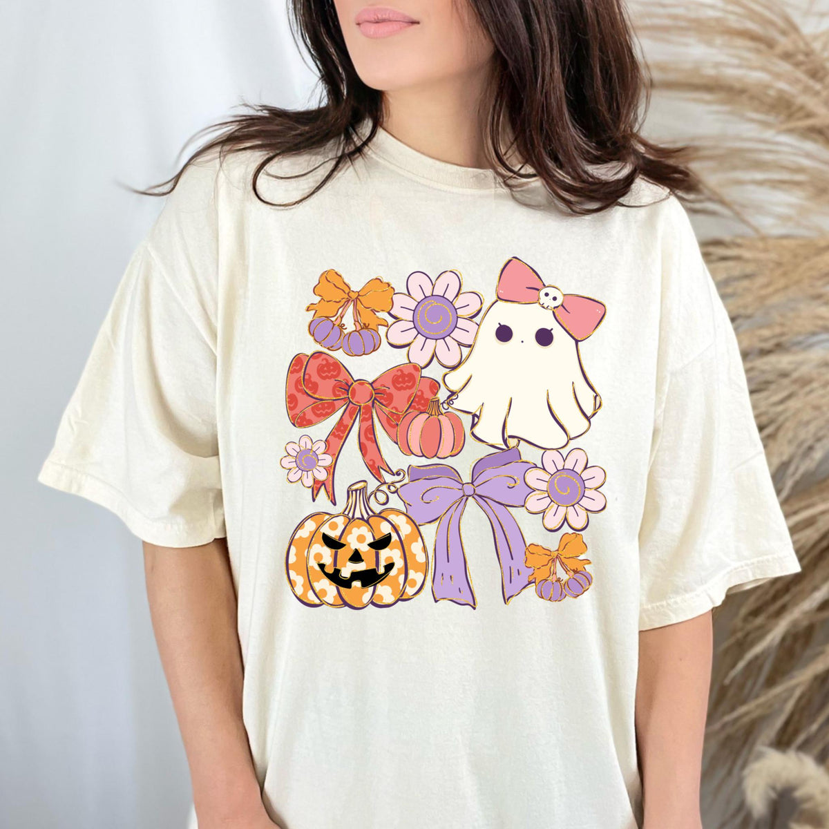 May include: Cream-colored t-shirt featuring a Halloween-themed graphic. The design includes a ghost with a pink bow, pumpkins, bows, and flowers in shades of orange, pink, and purple. The shirt has a relaxed fit.
