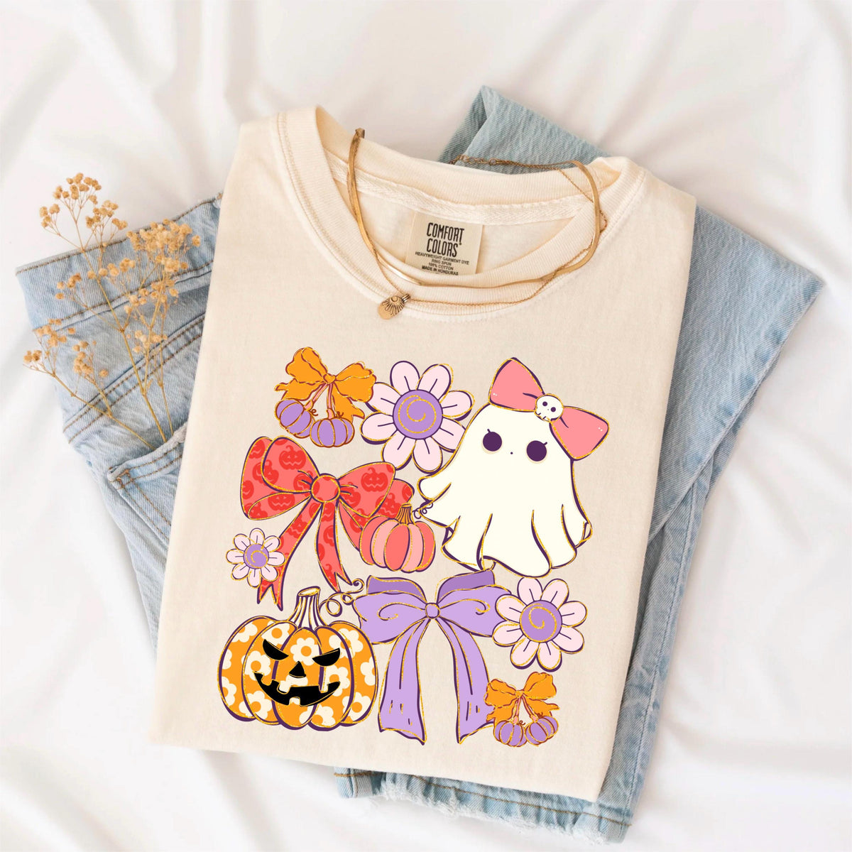 May include: Cream-colored Comfort Colors t-shirt featuring a Halloween-themed graphic. The design includes a ghost with a pink bow, pumpkins, bows, and flowers in shades of orange, pink, and purple. A gold necklace rests on the shirt.