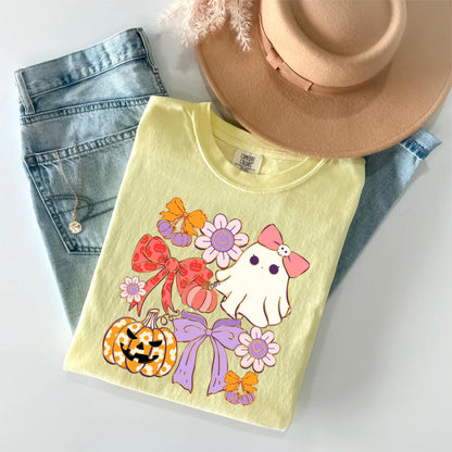 May include: A light yellow t-shirt featuring a Halloween-themed graphic with a ghost, bows, flowers, and pumpkins. The shirt is paired with light blue jeans and a tan hat. A necklace is visible in the jeans pocket.