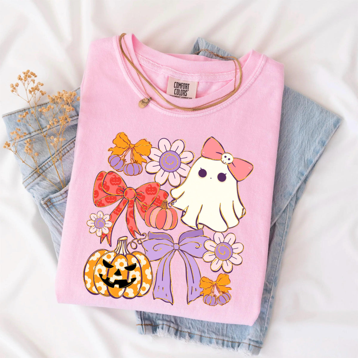 May include: A pink Comfort Colors t-shirt featuring a Halloween-themed graphic. The design includes a cute ghost with a pink bow, pumpkins, bows, and flowers. A gold necklace is draped over the shirt, which is folded on top of blue jeans.