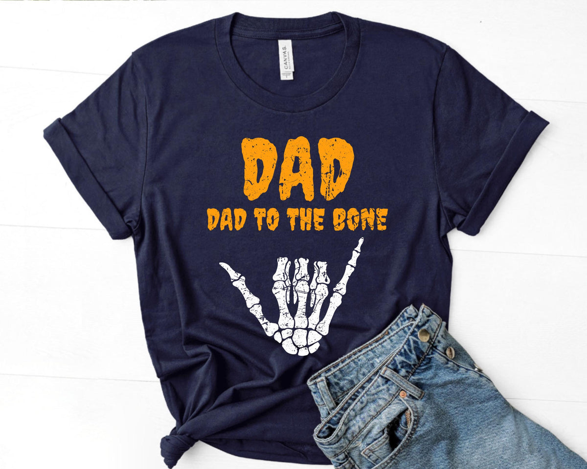 May include: Navy blue t-shirt with the text "DAD" in orange, and "DAD TO THE BONE" in orange. A white skeleton hand graphic is below the text. The shirt is paired with blue jeans.