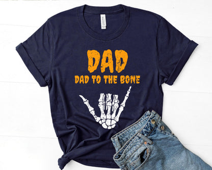 May include: Navy blue t-shirt with the text "DAD" in orange, and "DAD TO THE BONE" in orange. A white skeleton hand graphic is below the text. The shirt is paired with blue jeans.