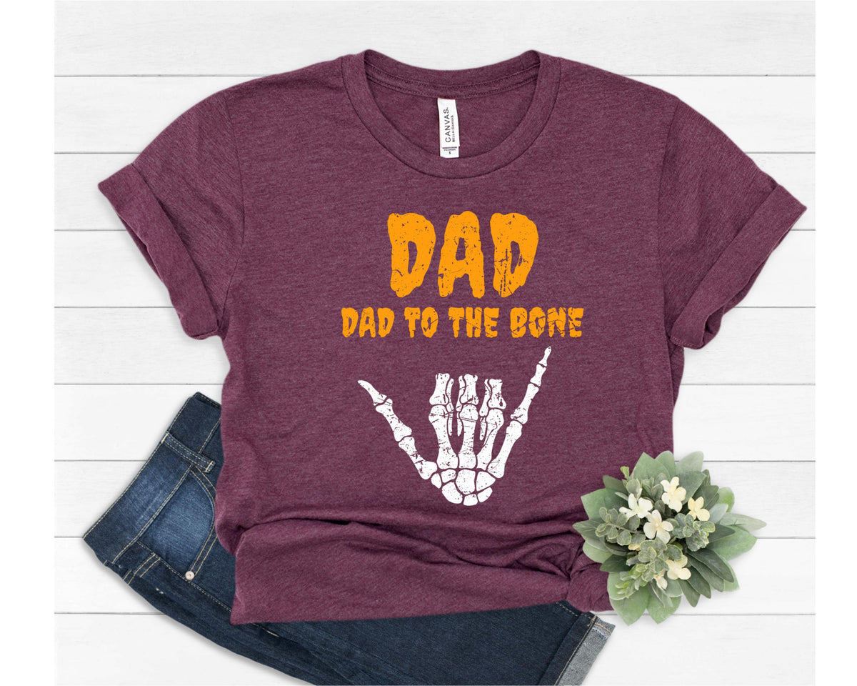 May include: Burgundy t-shirt with the text "DAD" in orange block letters above "DAD TO THE BONE". A white skeleton hand in the shaka sign is below the text. The shirt is paired with blue jeans and a small floral arrangement.