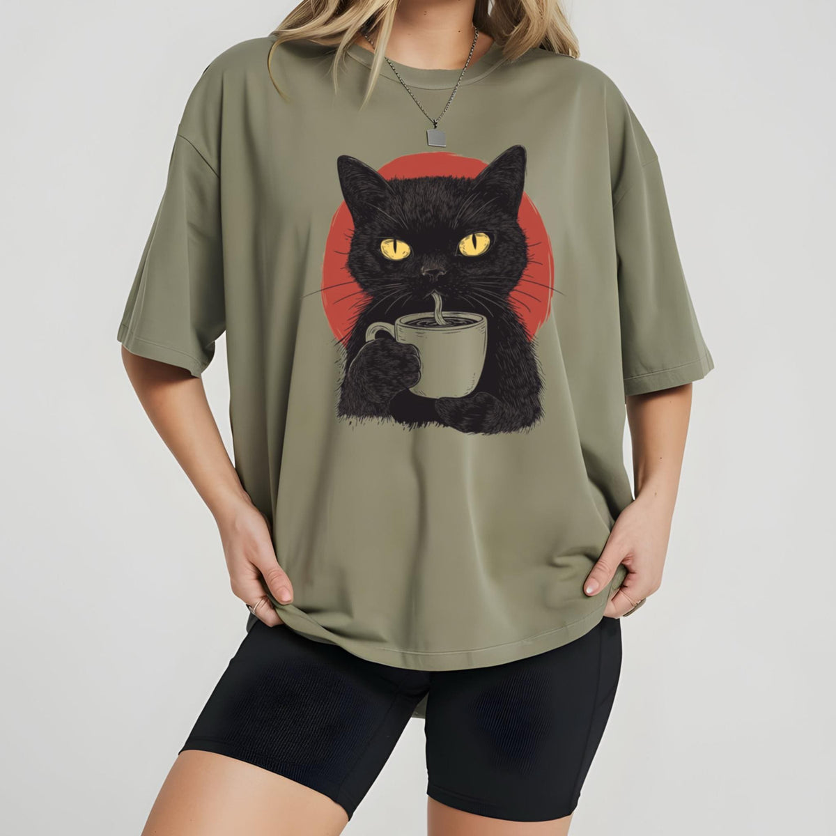 May include: An olive green t-shirt featuring a graphic illustration of a black cat with bright yellow eyes holding a light-colored mug. Behind the cat is a solid red circle. The shirt has short sleeves and a crew neckline, worn with black shorts.