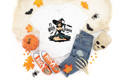 May include: White t-shirt with a graphic of a witch in a black dress and hat, with the text "It's Witch Season". The shirt is paired with blue jeans, orange sneakers, and Halloween decorations like a pumpkin and a skull.