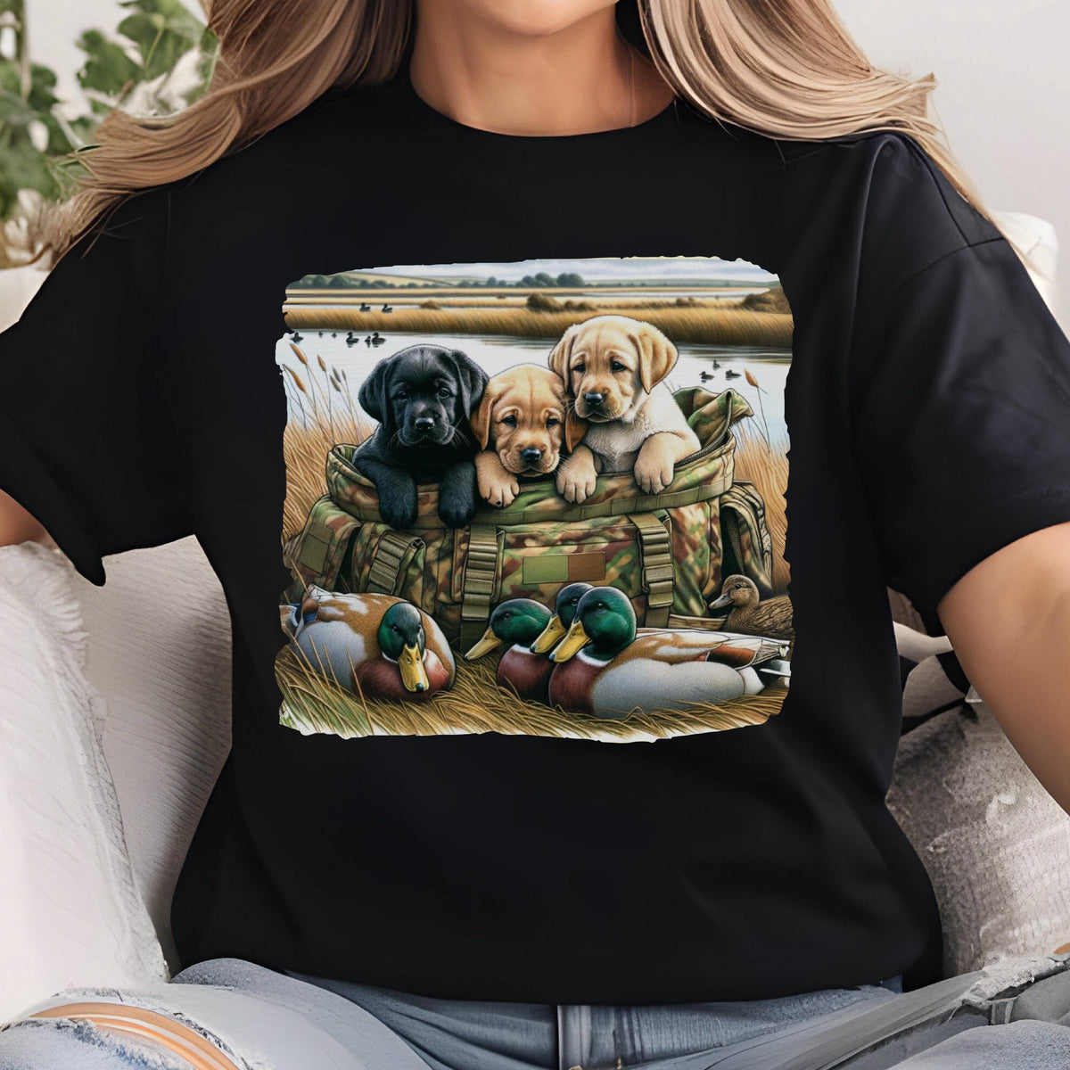 May include: Black t-shirt featuring a graphic of three Labrador puppies in a camouflage bag, with ducks in the foreground and a marsh scene in the background. The puppies are black, tan, and light yellow.