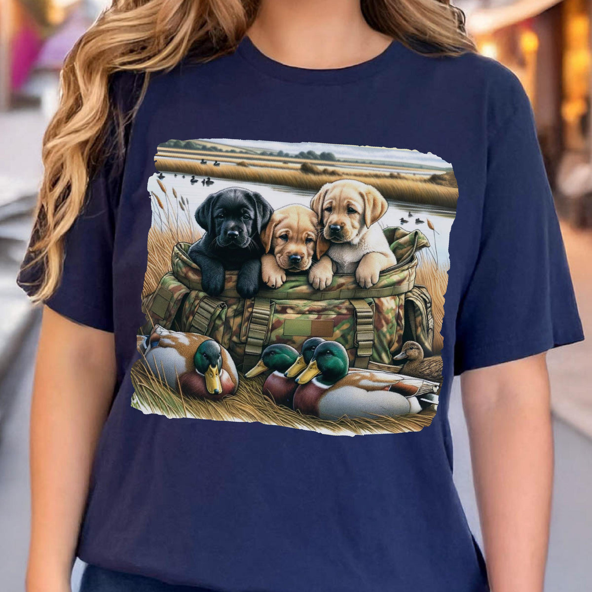 May include: Navy blue t-shirt featuring a graphic of three Labrador puppies in a camouflage bag, with ducks in the foreground and a marsh scene in the background. The puppies are black, tan, and light yellow. The ducks have green heads and brown bodies.
