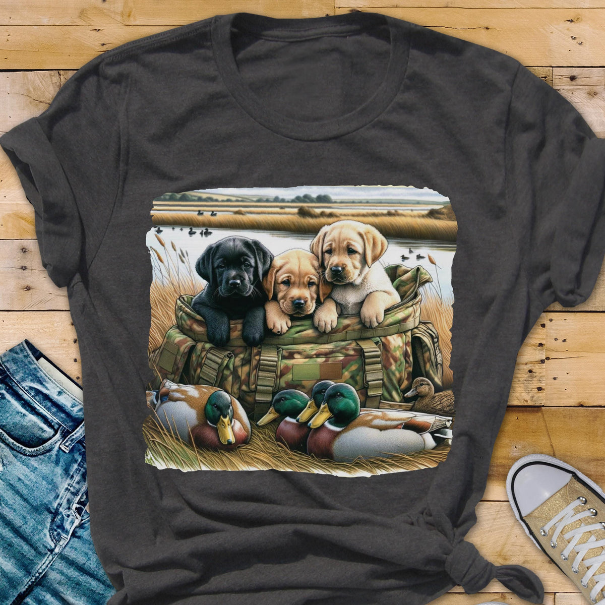 May include: Dark gray t-shirt featuring a graphic of three Labrador puppies in a camouflage bag, with ducks in the foreground and a marsh scene in the background. The puppies are black, tan, and cream-colored. The shirt is made of a soft material.
