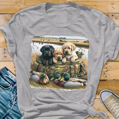 May include: A light gray t-shirt featuring a graphic of three Labrador puppies in a camouflage bag, with ducks in the foreground and a marsh scene in the background. The puppies are black, tan, and yellow. The shirt has a crew neck and short sleeves.