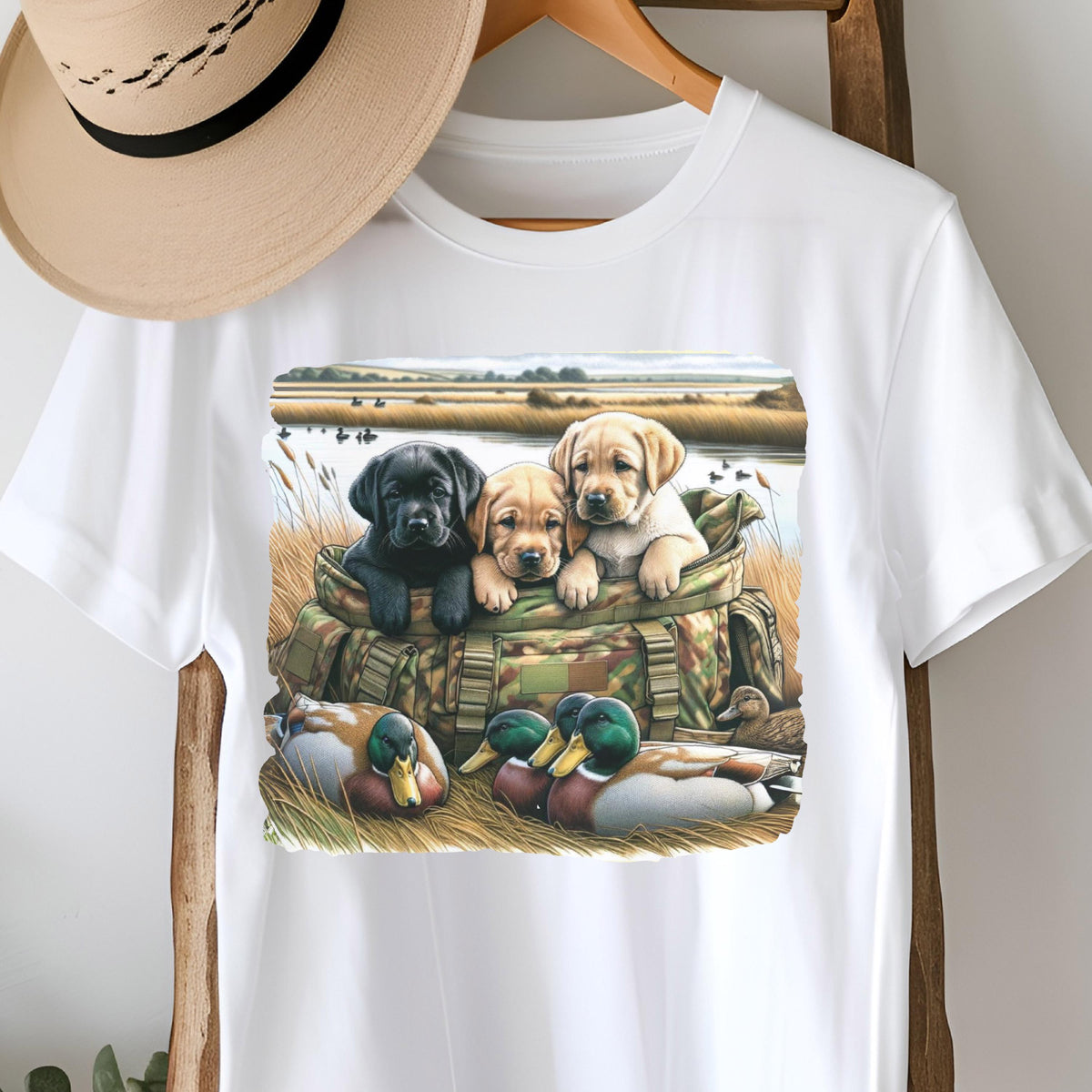 May include: White t-shirt featuring a graphic of three Labrador puppies in a camouflage bag, with ducks in the foreground and a marsh scene in the background. The puppies are black, tan, and yellow. A straw hat hangs above the shirt.