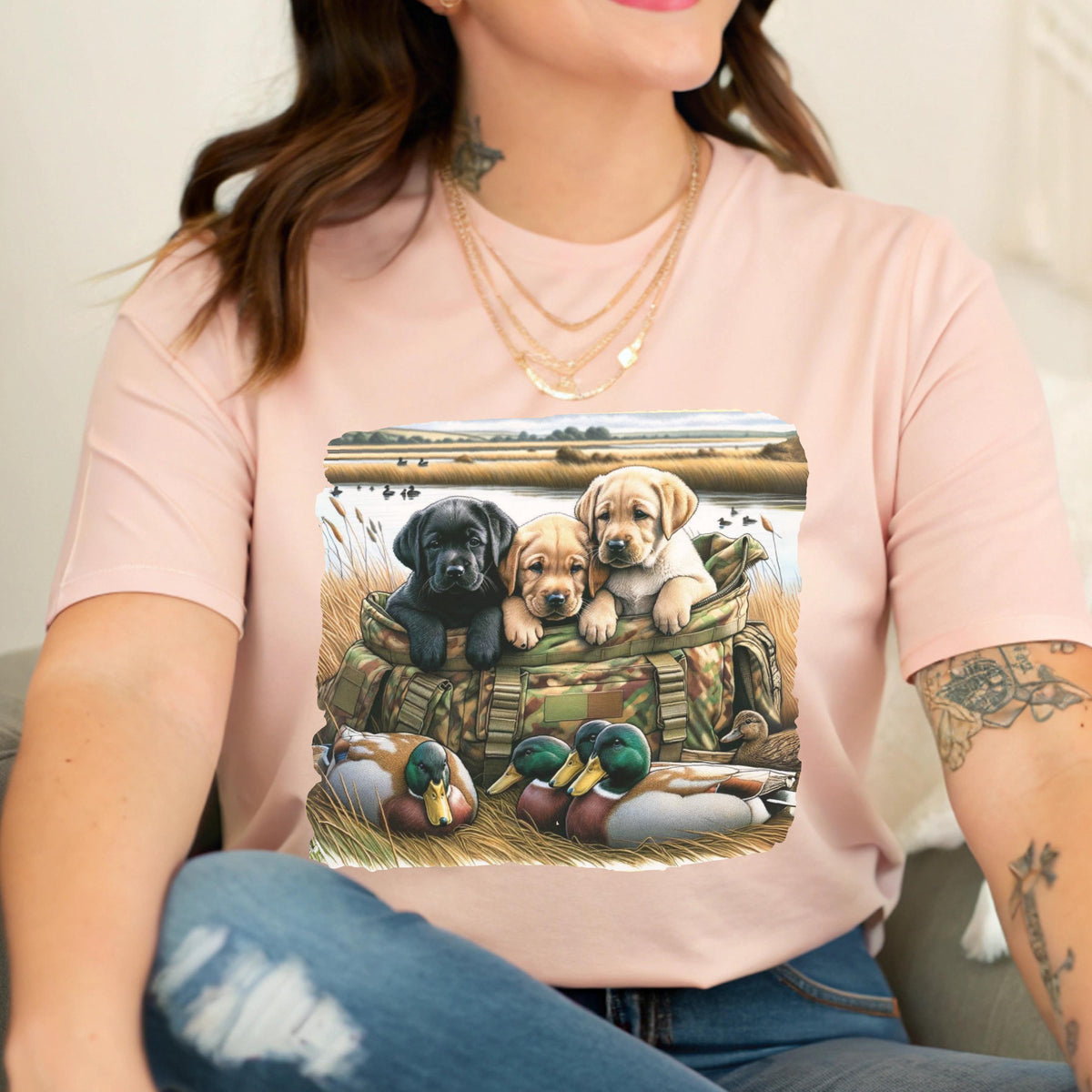 May include: Pink t-shirt featuring a graphic of three Labrador puppies in a camouflage bag, with ducks in the foreground. The puppies are black, tan, and yellow. The background depicts a marshy landscape.