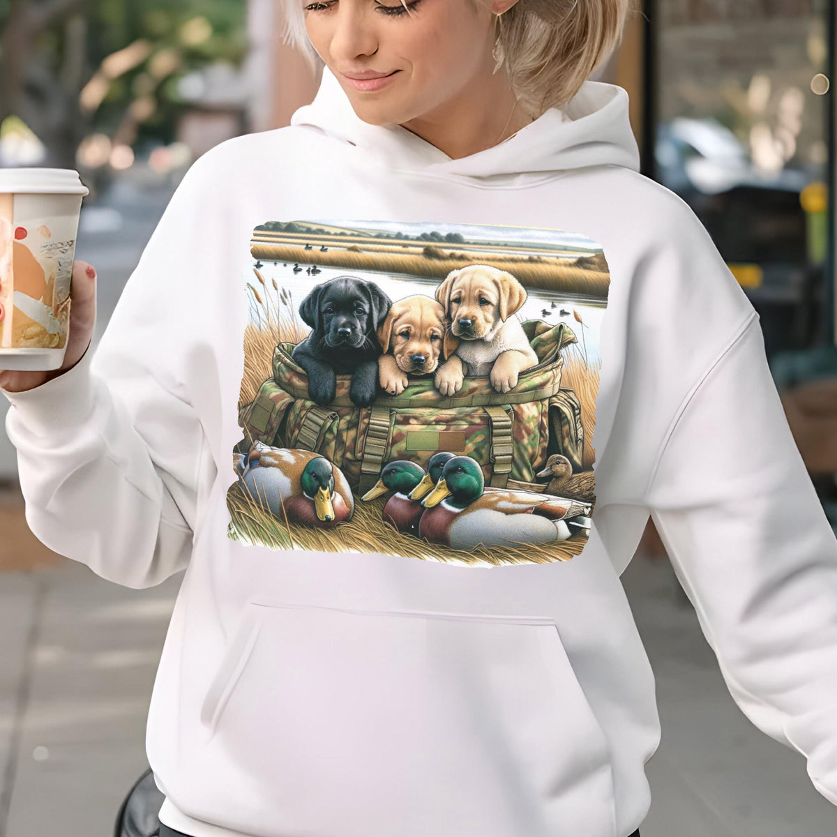 May include: A white hooded sweatshirt featuring a graphic print of three Labrador retriever puppies sitting inside a camouflage bag, with two mallard ducks resting in the foreground grass near a marshy landscape with water and reeds.