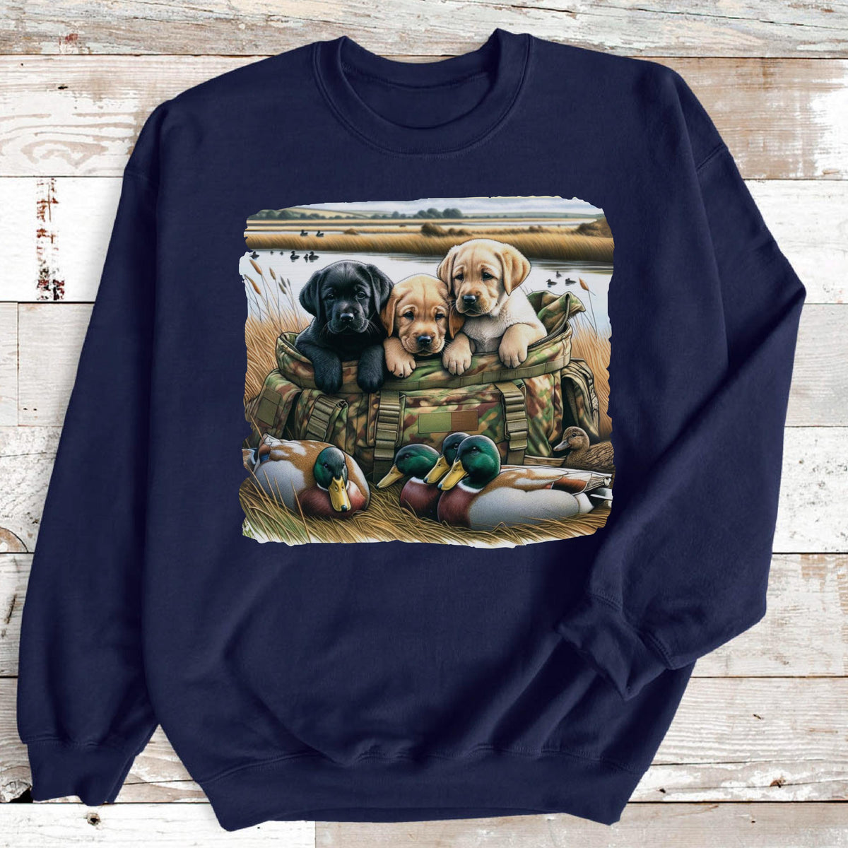 May include: A navy blue crewneck sweatshirt featuring a graphic print of three Labrador retriever puppies sitting inside a camouflage hunting bag, with two mallard ducks resting in the tall grass in the foreground near a body of water.