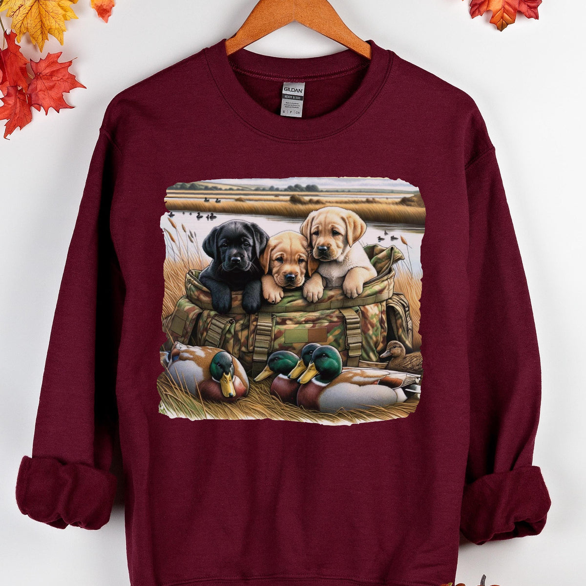 May include: A maroon long-sleeve sweatshirt featuring a graphic print of three Labrador retriever puppies sitting in a camouflage bag, with two mallard ducks resting in the foreground grass near a body of water and a field in the background.