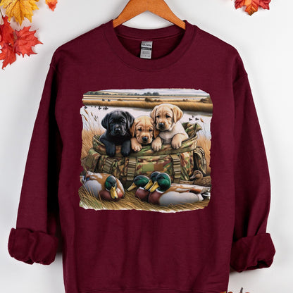 May include: A maroon long-sleeve sweatshirt featuring a graphic print of three Labrador retriever puppies sitting in a camouflage bag, with two mallard ducks resting in the foreground grass near a body of water and a field in the background.