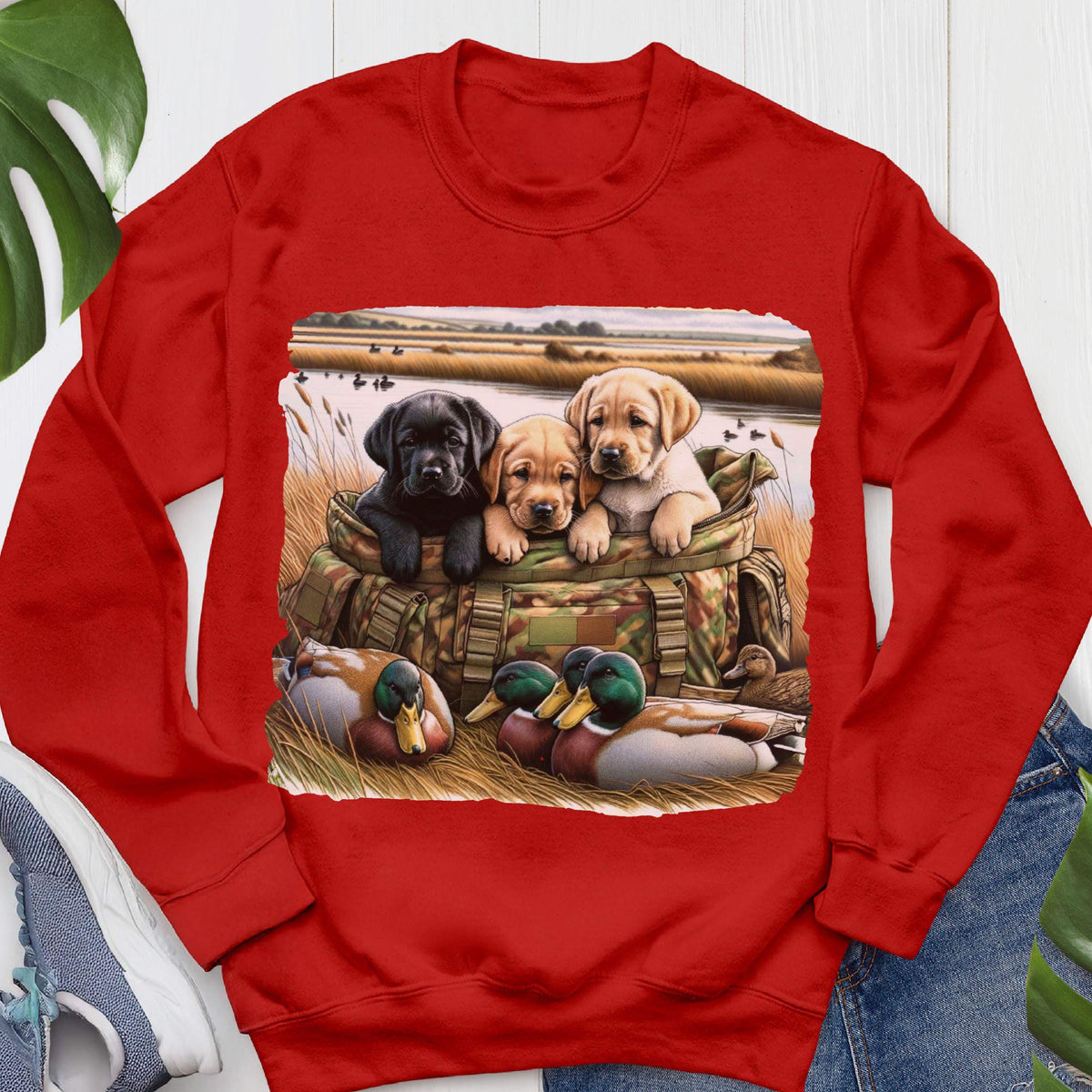 May include: A bright red crewneck sweatshirt featuring a central graphic print of three Labrador retriever puppies sitting inside a camouflage hunting bag, surrounded by mallard ducks in a grassy field near a body of water.