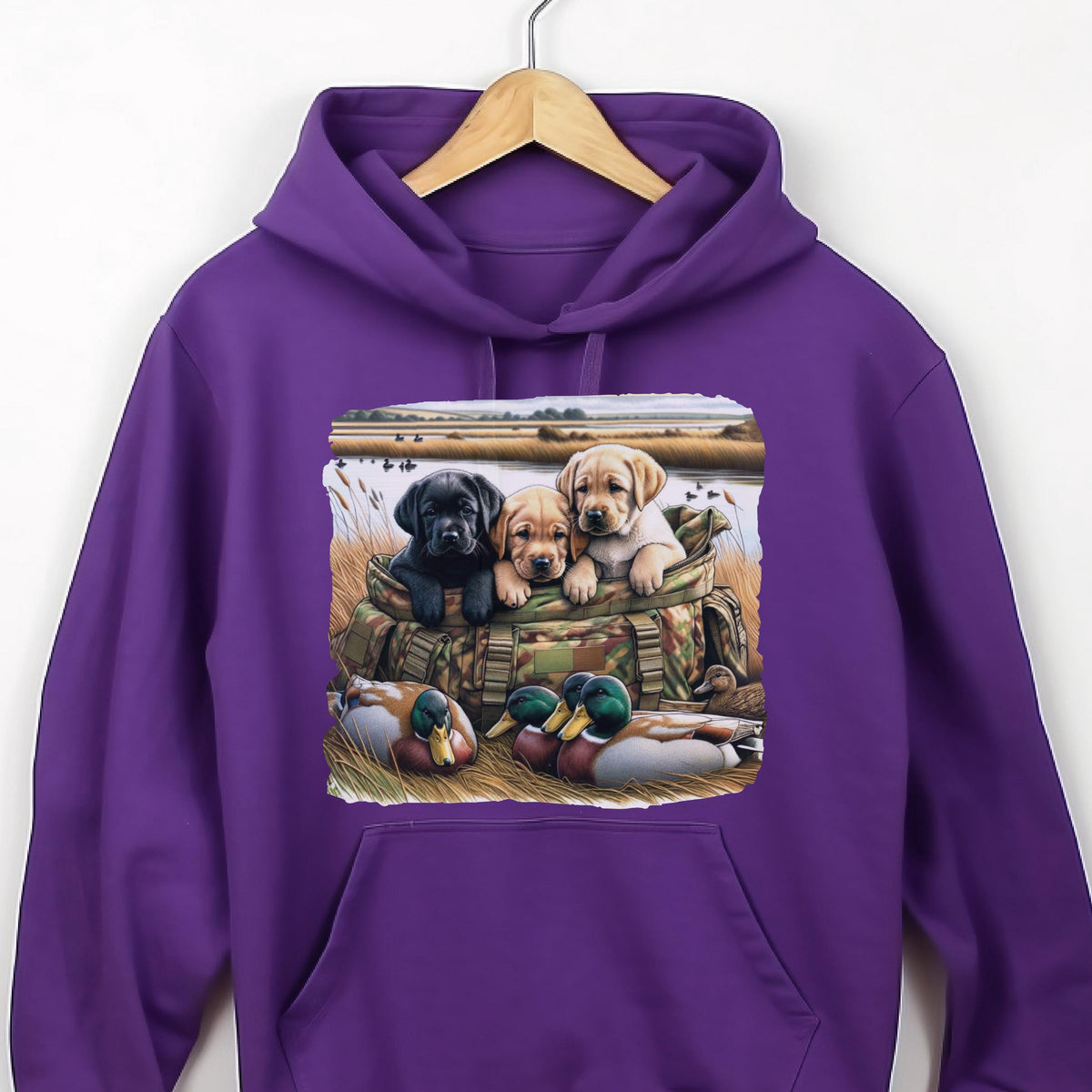 May include: A vibrant purple hooded sweatshirt featuring a graphic print of three Labrador puppies sitting inside a camouflage bag, with two mallard ducks resting in the foreground grass near a body of water and a field in the background.