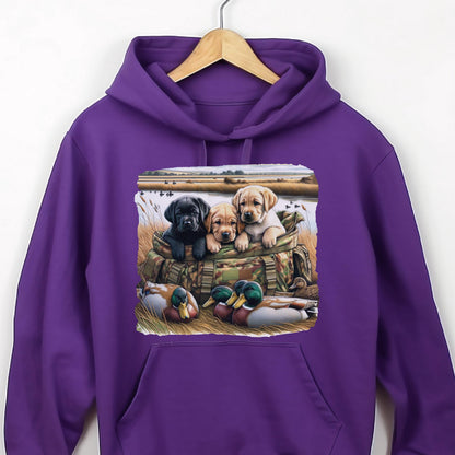 May include: A vibrant purple hooded sweatshirt featuring a graphic print of three Labrador puppies sitting inside a camouflage bag, with two mallard ducks resting in the foreground grass near a body of water and a field in the background.