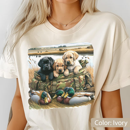 May include: An ivory t-shirt featuring a graphic print of three Labrador retriever puppies sitting inside a camouflage duffel bag, surrounded by mallard ducks in a grassy field with a pond and reeds in the background.