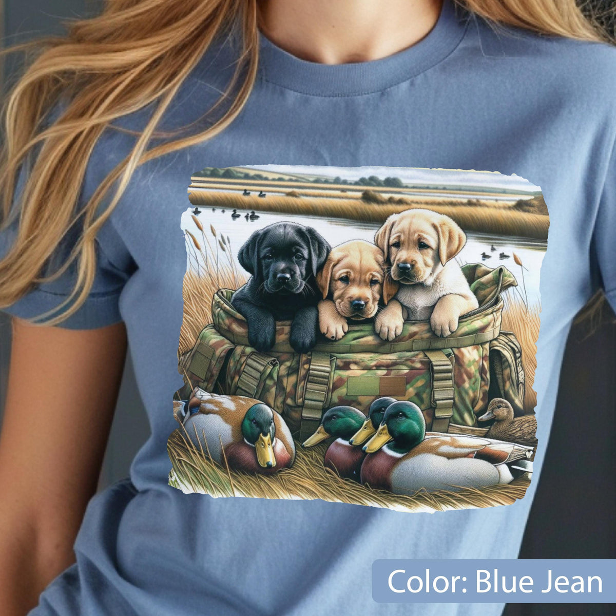 May include: A blue jean colored t-shirt featuring a graphic print of three Labrador retriever puppies, one black and two yellow, sitting inside a camouflage patterned bag, with two mallard ducks resting in the grass in the foreground.