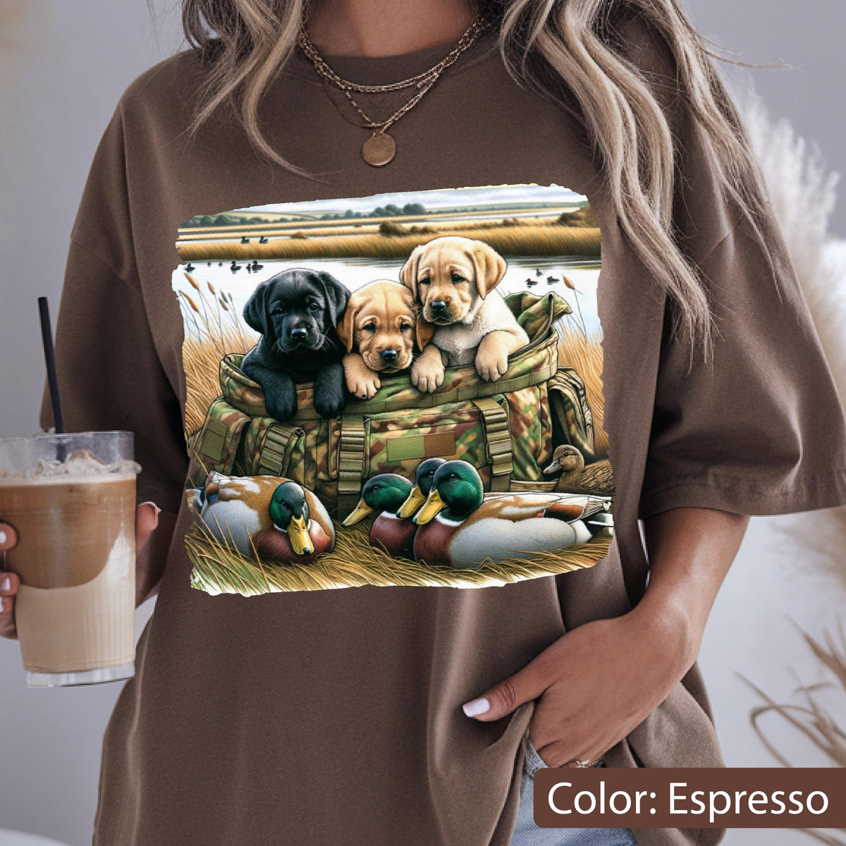 May include: An espresso-colored t-shirt featuring a graphic print of three puppies, one black and two yellow, sitting inside a camouflage-patterned bag. In the foreground, two mallard ducks rest in tall grass near a body of water and reeds.