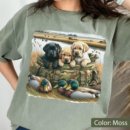 May include: A moss green t-shirt featuring a graphic print of three Labrador retriever puppies sitting inside a camouflage hunting bag, with two mallard ducks resting in the tall grass in the foreground and a marshy landscape in the background.