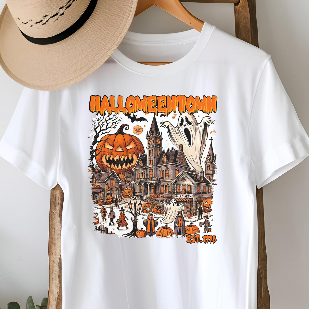 May include: White t-shirt featuring a Halloween-themed graphic. The design includes a jack-o'-lantern, a haunted house, ghosts, and the text "Halloweentown" in orange. The shirt also has the text "EST. 1998".