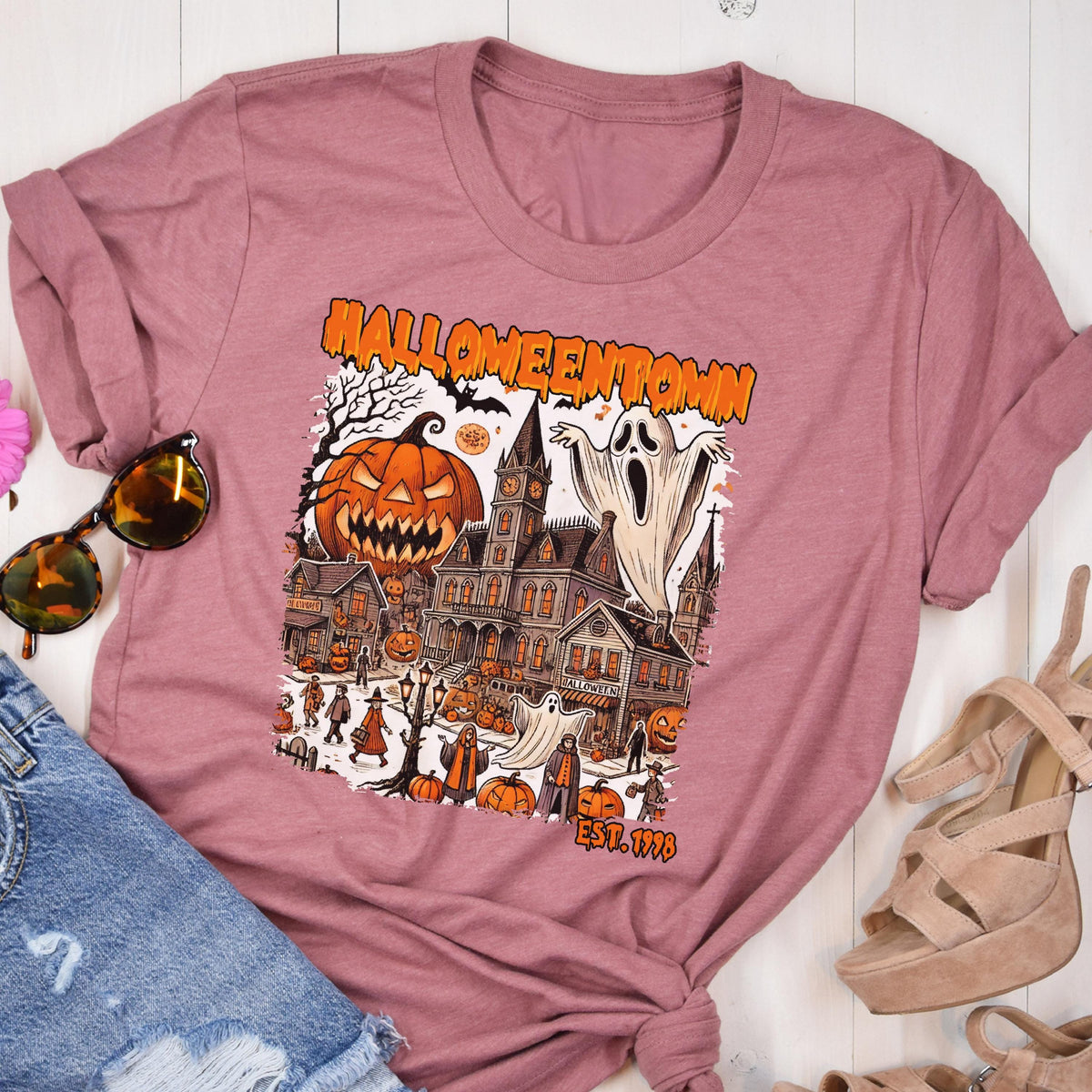 May include: Dusty rose t-shirt featuring a Halloween-themed graphic. The design includes a haunted house, jack-o'-lanterns, ghosts, and the text "Halloweentown" and "EST. 1998" in orange. The shirt is folded, with sunglasses and a pair of sandals visible.