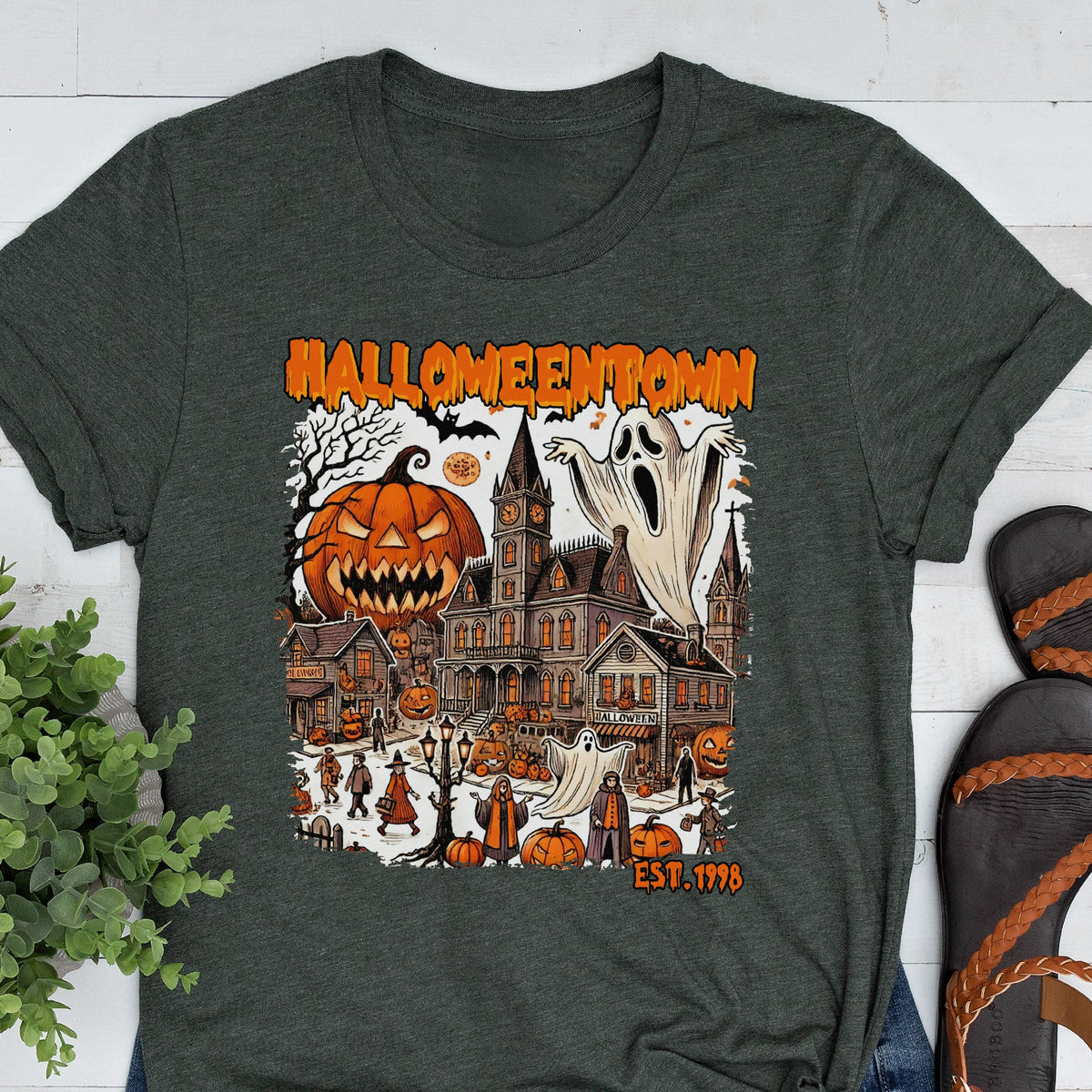 May include: Dark gray t-shirt featuring a Halloween-themed graphic. The design includes a jack-o'-lantern, a haunted town, ghosts, and the text "Halloweentown" and "EST. 1998" in orange. The shirt has short sleeves and a crew neck.