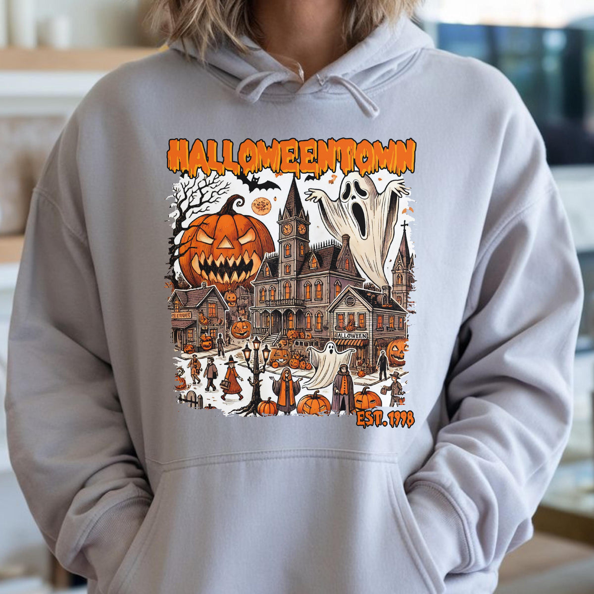 May include: A light gray hooded sweatshirt featuring a graphic print of a town scene with the text HALLOWEENTOWN EST. 1998. The illustration includes a large jack-o'-lantern, a tall clock tower building, ghosts, and various figures in a town square.