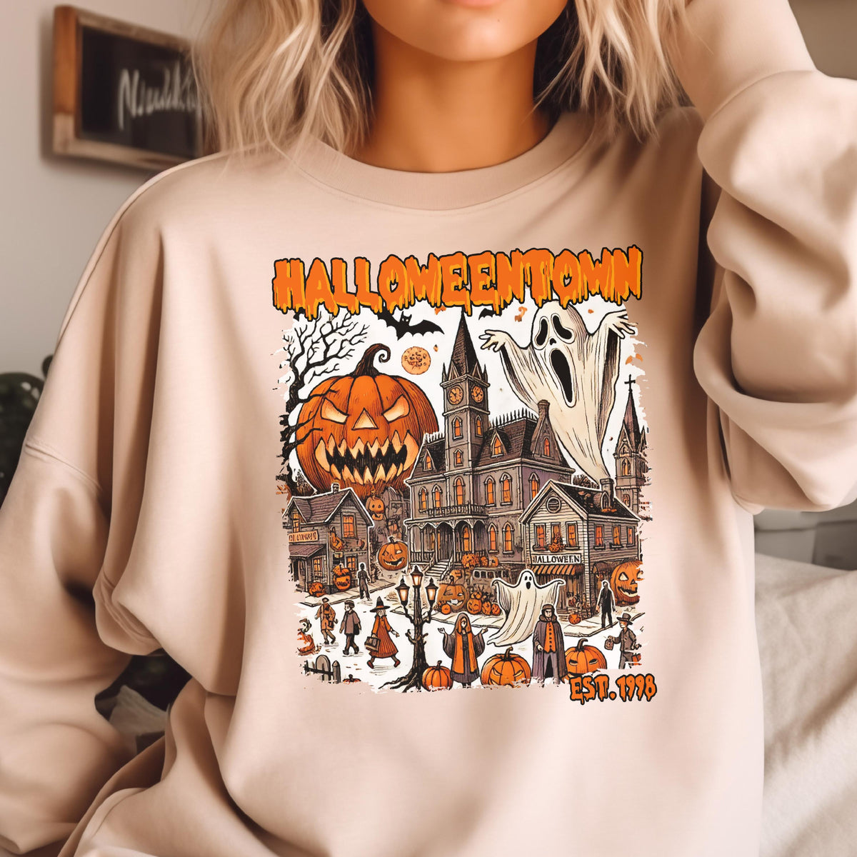 May include: A beige crewneck sweatshirt featuring a graphic print with the text HALLOWEENTOWN EST. 1998. The illustration depicts a spooky town scene with a large jack-o'-lantern, ghosts, a clock tower, and various figures in a Halloween setting.