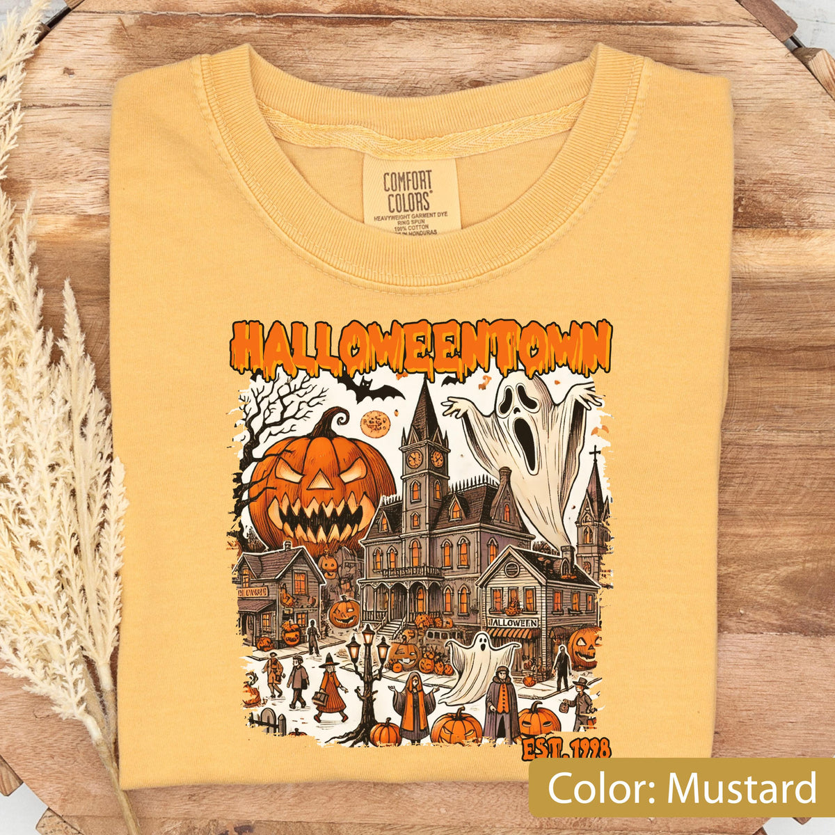May include: A mustard yellow t-shirt featuring a graphic print with the text "HALLOWEENTOWN" and "EST. 1998". The illustration depicts a spooky town scene with a large jack-o'-lantern, a haunted mansion, ghosts, and various figures in costumes.