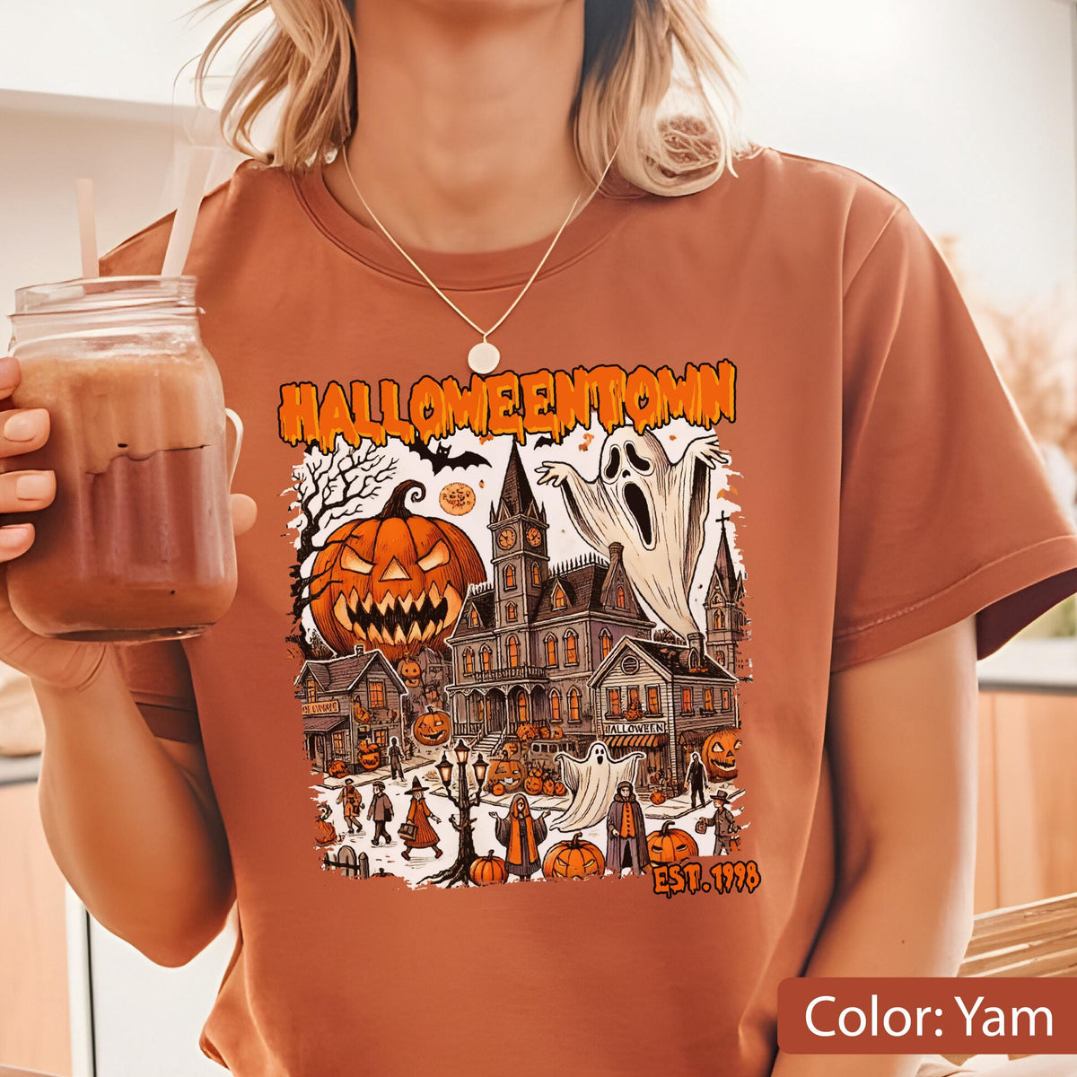 May include: A yam-colored t-shirt featuring a graphic print with the text HALLOWEENTOWN and EST. 1998. The illustration depicts a spooky town scene with a large jack-o'-lantern, a haunted clock tower, ghosts, and various figures in costumes.