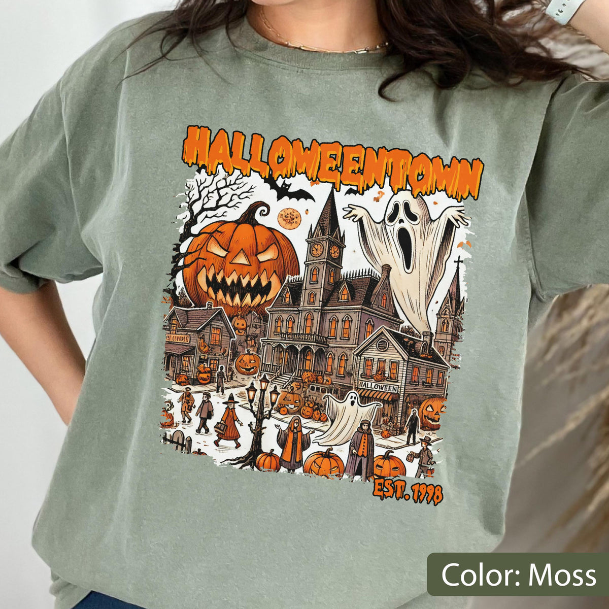May include: A moss green t-shirt featuring a graphic print of a town scene with the text HALLOWEENTOWN and EST. 1998. The illustration includes a large jack-o'-lantern, a tall clock tower, multiple houses, ghosts, and figures in costumes.