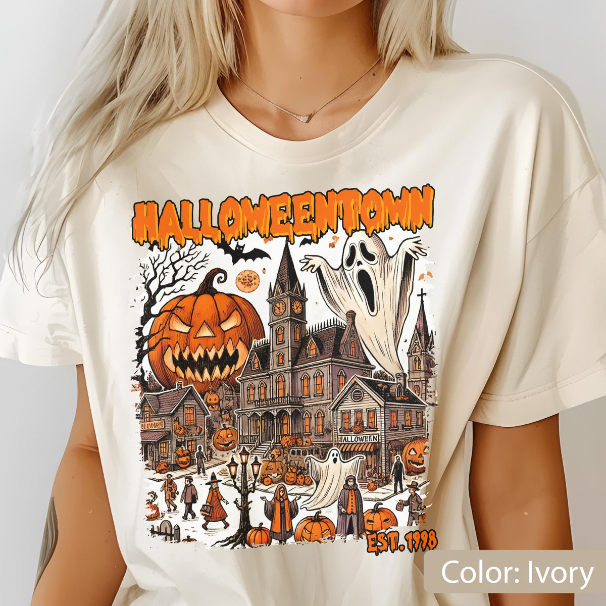 May include: An ivory t-shirt featuring a graphic print with the text HALLOWEENTOWN EST. 1998. The design includes a large jack-o'-lantern, a haunted mansion, ghosts, bare trees, and small figures in a town setting with orange and black accents.