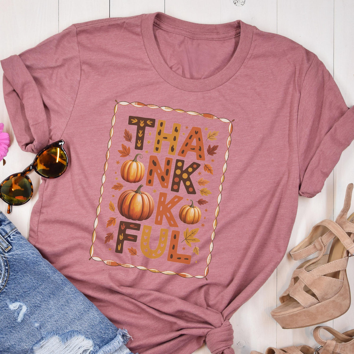 May include: A mauve short-sleeve t-shirt featuring a rectangular frame containing the text THANKFUL in brown and orange block letters, interspersed with illustrations of orange pumpkins and scattered autumn leaves in shades of yellow and brown.