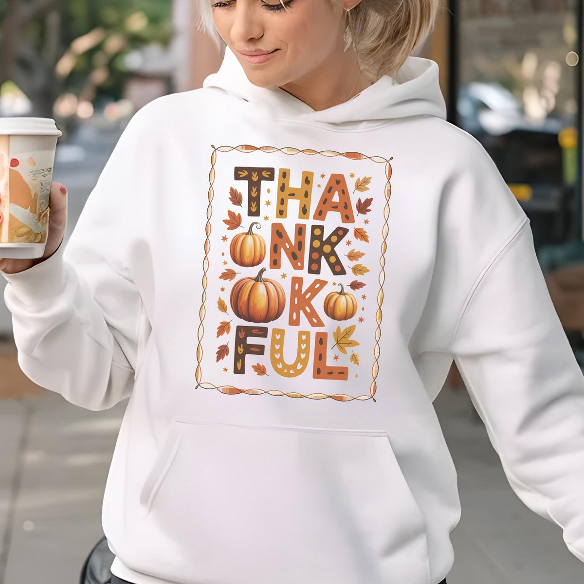 May include: White hooded sweatshirt with a "THANKFUL" graphic in fall colors. The design includes pumpkins and autumn leaves. The sweatshirt has a front pocket and a drawstring hood. A person is holding a coffee cup.