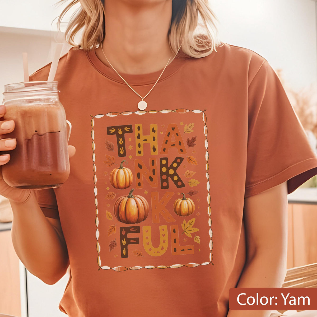 May include: A yam-colored t-shirt with a fall-themed graphic. The graphic features the word "THANKFUL" with pumpkins and autumn leaves. The design is framed with a dotted border. A glass jar with a straw is visible.