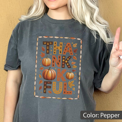 May include: Pepper-colored t-shirt with the word "THANKFUL" in a fall-themed design. The design includes pumpkins and autumn leaves. The shirt is a comfortable, casual style.