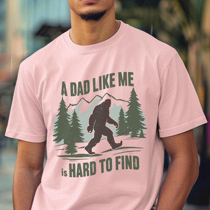 May include: A light pink short-sleeve t-shirt featuring a dark green graphic of a Bigfoot silhouette walking through a forest with mountains in the background. The text reads A DAD LIKE ME is HARD TO FIND in a bold, dark green sans-serif font.
