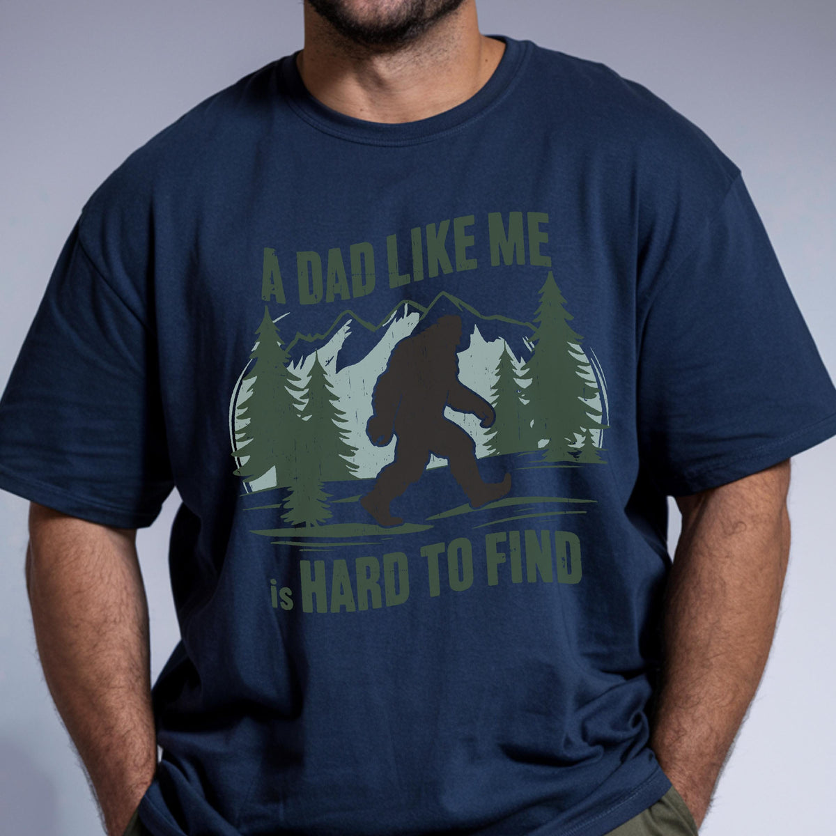 May include: A navy blue short-sleeved t-shirt featuring a graphic print of a dark silhouette of Bigfoot walking through a forest with mountains in the background. The text reads A DAD LIKE ME is HARD TO FIND in olive green lettering.