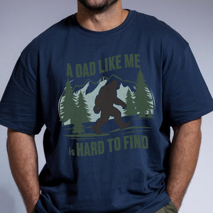 May include: A navy blue short-sleeved t-shirt featuring a graphic print of a dark silhouette of Bigfoot walking through a forest with mountains in the background. The text reads A DAD LIKE ME is HARD TO FIND in olive green lettering.