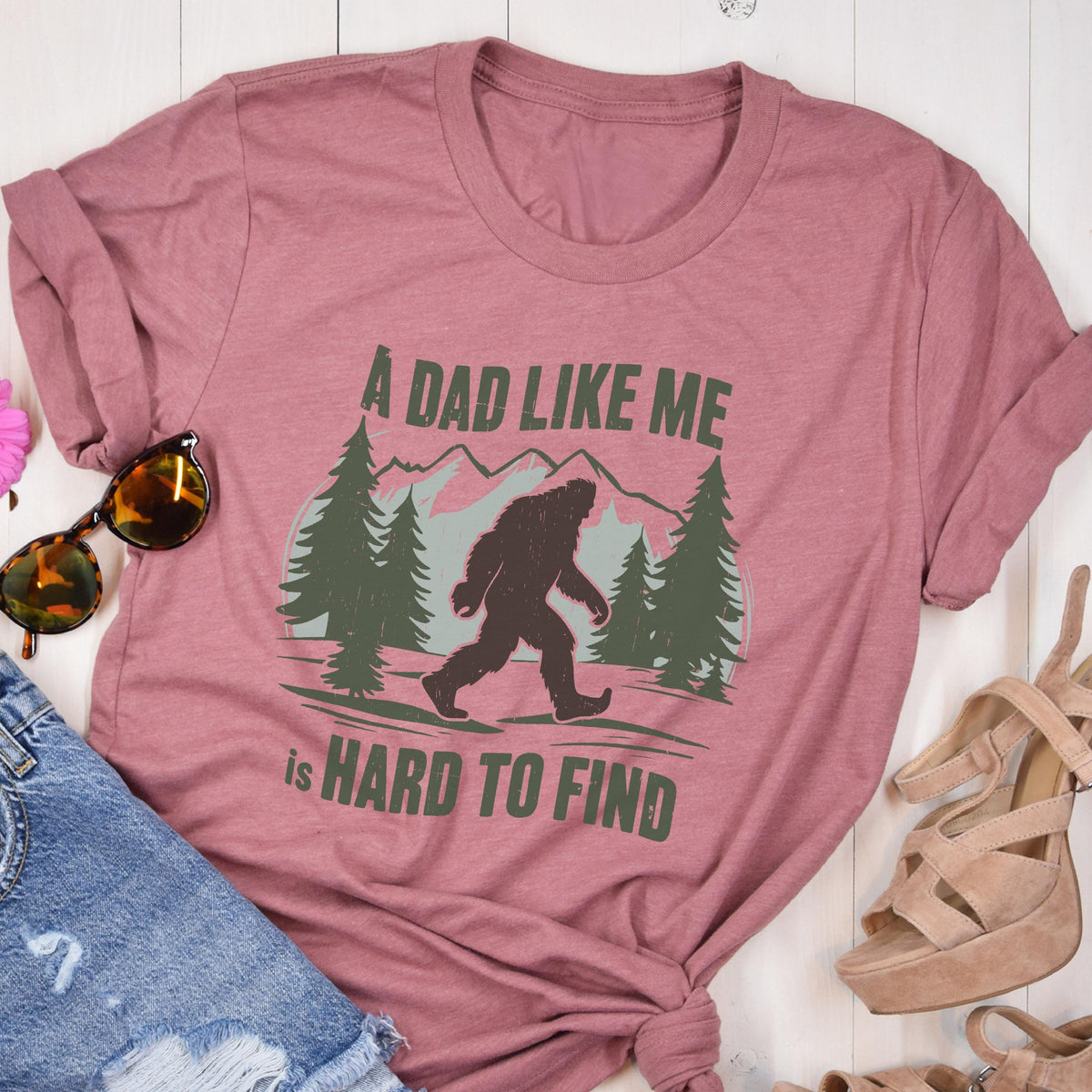 May include: A mauve short-sleeve t-shirt featuring a dark green graphic of a Bigfoot silhouette walking through a forest with mountains in the background. The text "A DAD LIKE ME is HARD TO FIND" is printed in dark green capital letters around the graphic.