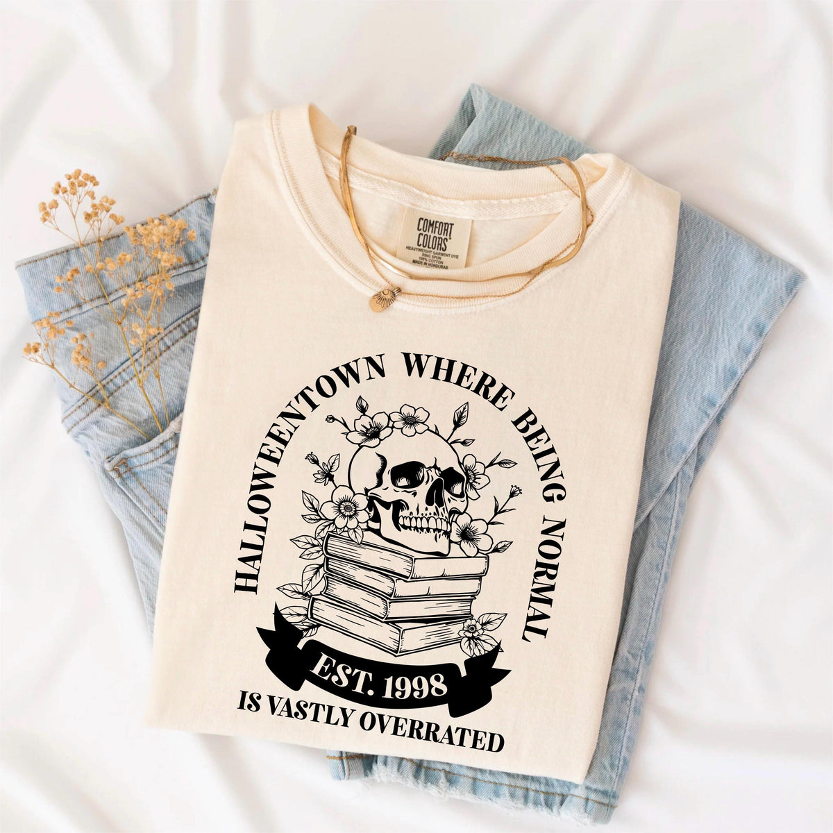May include: Cream-colored t-shirt with a black graphic featuring a skull adorned with flowers atop a stack of books. The text reads "Halloweentown Where Being Normal Is Vastly Overrated" and "Est. 1998". A gold necklace rests on the shirt.