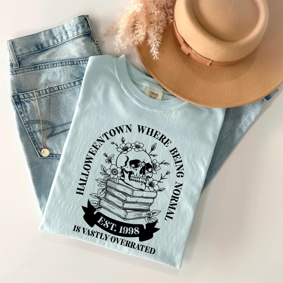 May include: A light blue t-shirt with a graphic that reads "Halloweentown where being normal is vastly overrated" with a skull, flowers, and books. The shirt is paired with blue jeans and a tan hat.