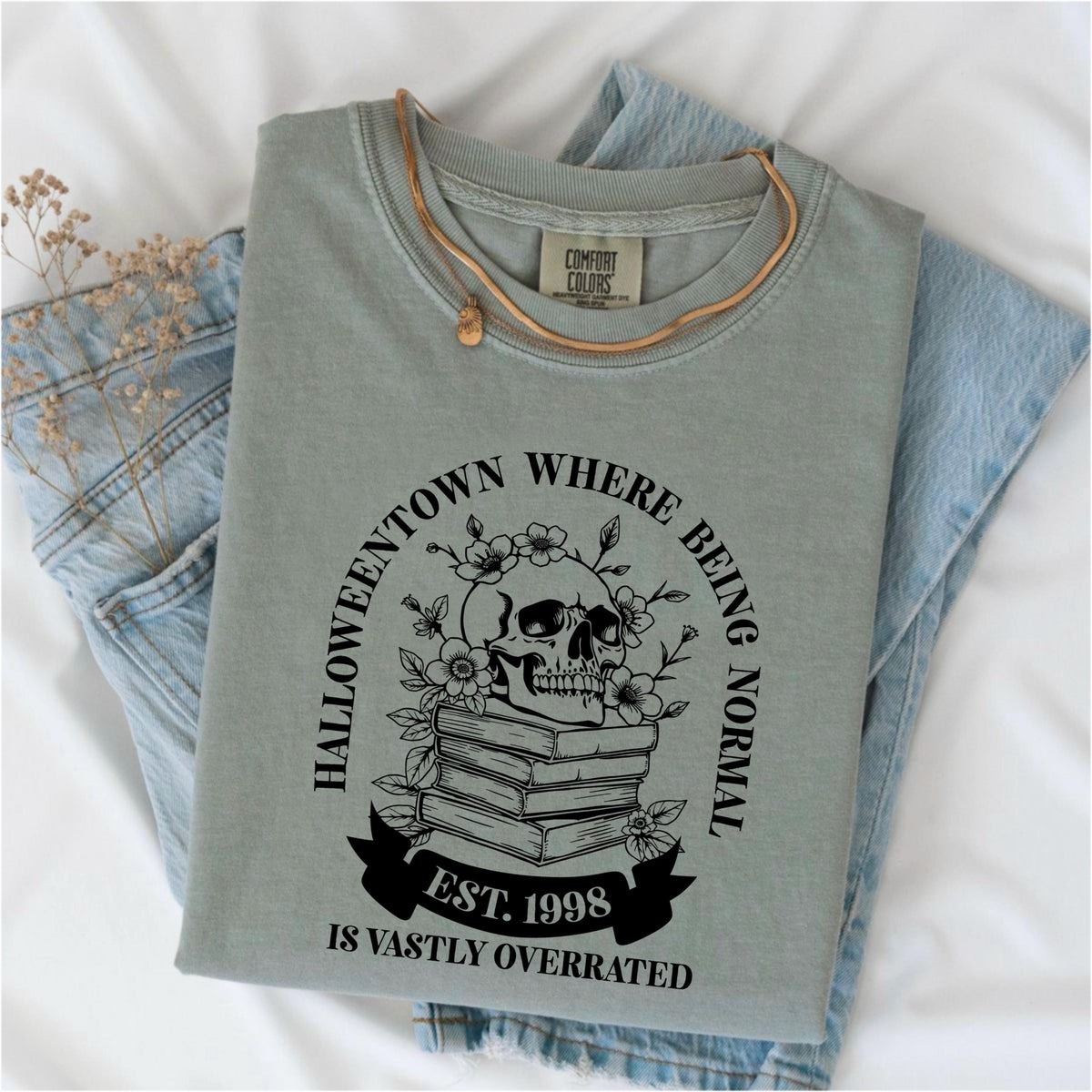 May include: Sage green Comfort Colors t-shirt with a graphic featuring a skull, flowers, and books. The text reads "Halloweentown where being normal is vastly overrated. Est. 1998." A gold necklace rests on the shirt, which is folded on denim.