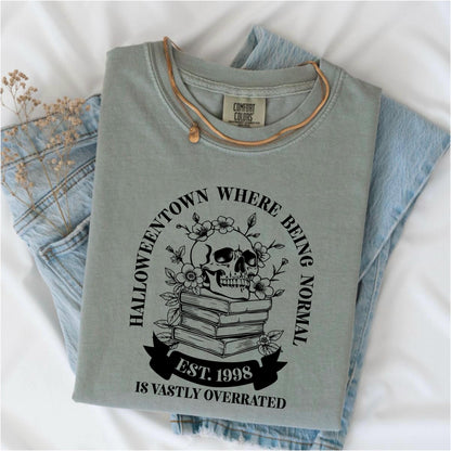 May include: Sage green Comfort Colors t-shirt with a graphic featuring a skull, flowers, and books. The text reads "Halloweentown where being normal is vastly overrated. Est. 1998." A gold necklace rests on the shirt, which is folded on denim.