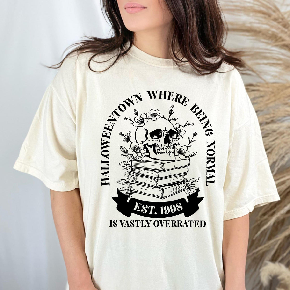 May include: Cream-colored t-shirt featuring a black and white graphic. The graphic includes a skull atop stacked books, surrounded by flowers, and the text "Halloweentown Where Being Normal Is Vastly Overrated. Est. 1998."