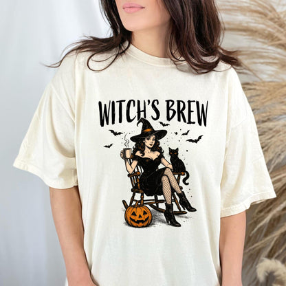May include: A cream-colored t-shirt featuring a graphic print with the text "WITCH'S BREW" above an illustration of a person in a black dress and hat sitting in a rocking chair with a black cat, a steaming mug, and a carved orange pumpkin.