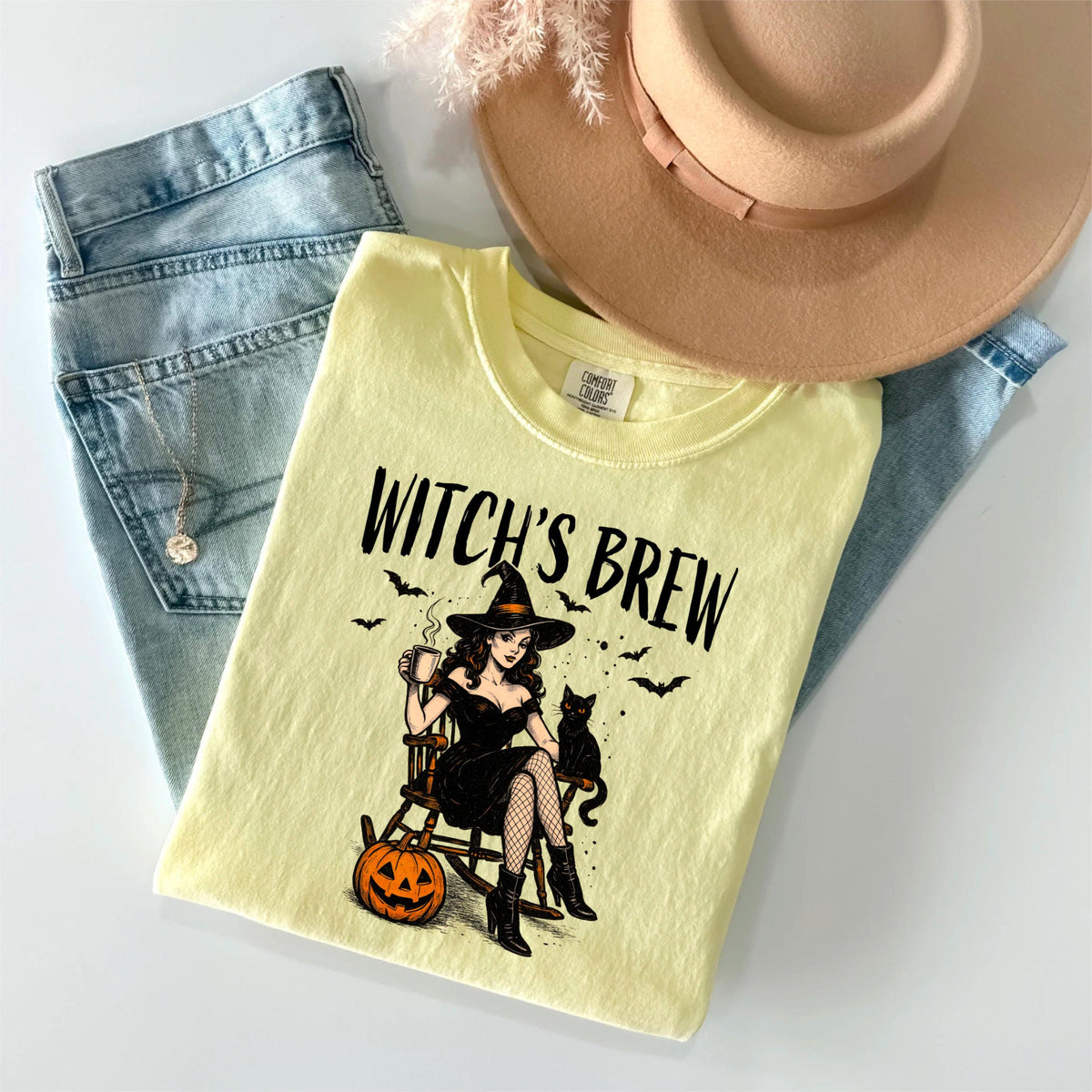 May include: A pale yellow t-shirt featuring a graphic of a person in a black dress and witch hat sitting in a rocking chair with a black cat, a pumpkin, and bats. The text "WITCH'S BREW" is printed above the illustration in a black font.